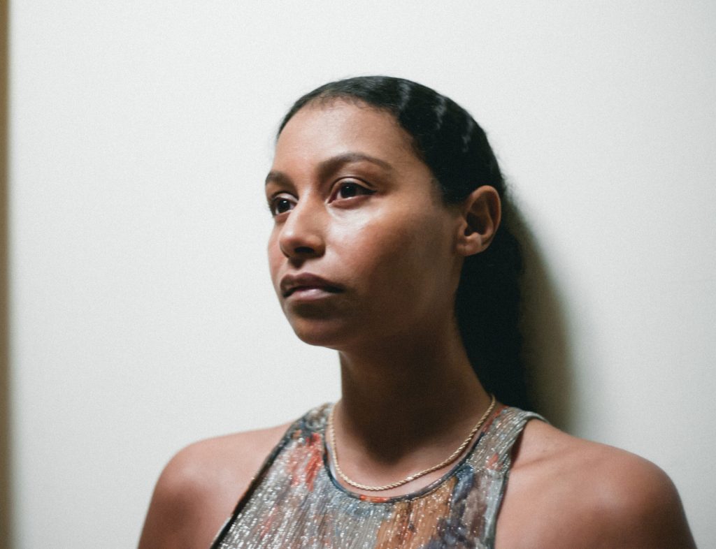 Photo of Giada Scodellaro. Her dark wavy hair trails behind her ear, smooth and shiny. She stands against a white wall, looking off beyond the camera. She wears a sparkling sleeveless multicolored abstract shirt and a delicate but sturdy necklace. Her dark eyes command the image and her brown skin glows in light and shadow. 