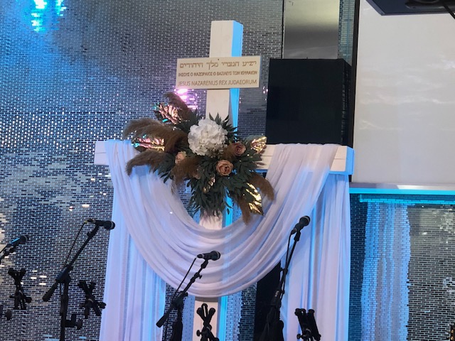A photo from the author of a cross draped in lilac fabric with writing in Hebrew, Ukrainian and Latin. THe cross is decorated with flowers and leaves, and microphones stand before it, waiting for speakers. The back wall is shiny, mirrored mosaics. 