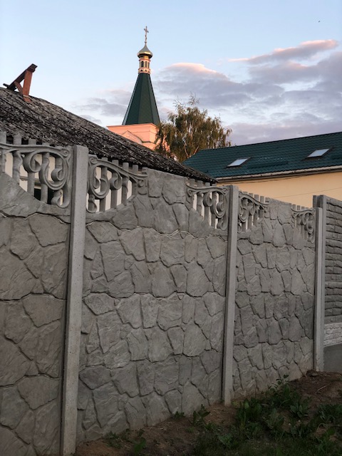 A photo from the author of a fence made to look like a stone wall, behind which are yellow walls, a sloped black roof, and the spire of a church, topped with a fancy, shiny dome and a giant cross. 