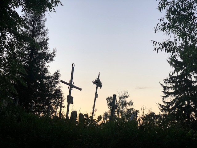 Author photograph. Crosses silhouetted at sunset before a sky that is purple/light blue by the treetops but yellow orange where it meets the earth. 