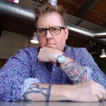 Jason O'Toole poses at a table, with one arm resting on its surface and the other hand up against his chin. He wears a blue shirt with rolled up sleeves, displaying his tattoos, and a quizzical expression.  
