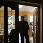 Edward Lee, photographed in silhouette in a dark room with wood and glass doors. Behind him, through the doorway is a brighter room with light wood and white paint. 