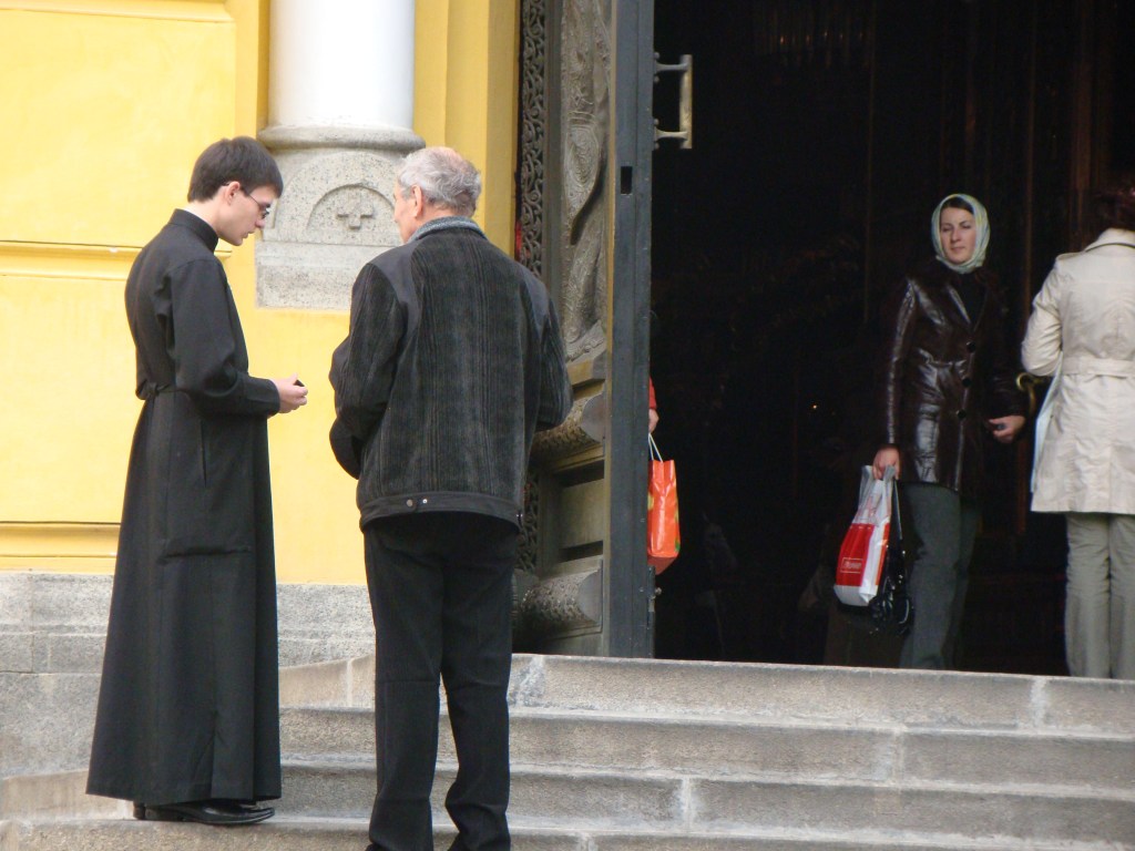 A photograph of Saint Vladimir Cathedral in Kyiv, circa 2008 by VasenkaPhotogrpahy. A man in long black robes speaks to an older man in a black jacket on the steps, standing in front of the cathedral's yellow external walls. Inside a woman in a grey headscarf looks through the large ornate cathedral doors from the shadowy entryway. She wears a shiny jacket and holds a shopping bag. Next to her a woman faces into the cathedral, wearing grey/green pants and a cream jacket.