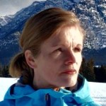 Laura Swart stands in blue before a mountain. Her face is half in sun, half in shadow, and her dark blond hair in a neat short ponytail. 