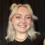 Standing against a black background, Theo Bee smiles brightly with his shoulder length blond hair partially pinned up. He wears a black shirt and a small necklace. 
