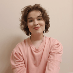 Author Photo: Klein Voorhees sits before a cream wall. Their hair is shorter than shoulder length, curly brown. They smile, in a large pink sweater and dangly cream earrings.