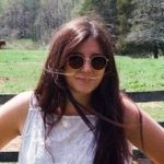 Artist photo: Giuliana Eggleston stands before a wooden fence and a field, in sunglasses. She wears a white tank top and her long brown hair frames her face.