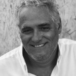 A black and white photograph of João Luís Barreto Guimarães, smiling at the camera, his white shirt standing out against the grass behind him, his hair standing up in short, spiky waves.