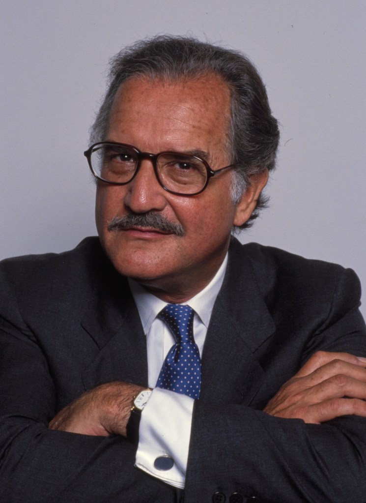 Photograph of Carlos Fuentes: he has a skeptical look to his friendly eyes, and a smile under his graying mustache. He wears rounded plastic frame glasses and a dark suit, with a blue tie. He crosses his arms and looks into the camera. 