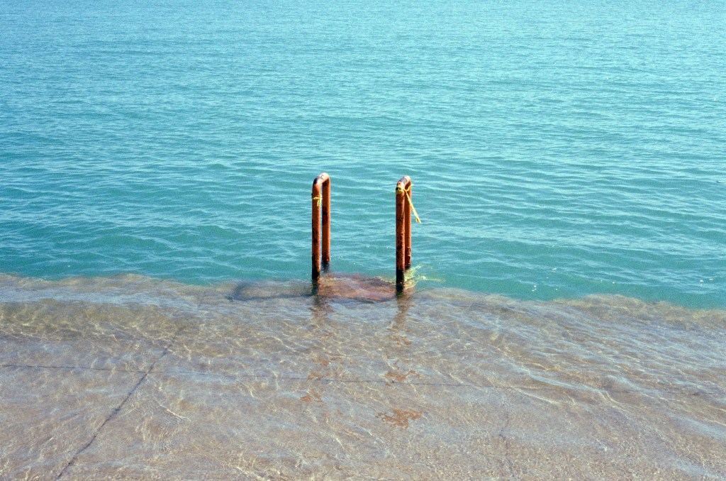 Art description: Summer in Chicago 2 by Giuliana Eggleston shows the edge of the city, where the cement along the lake is covered with water, wavering and refracting sun and shadow on the ground until it stops and the lake officially begins, a border marked by the rusty supports of a ladder down. The lake extends beyond the edges of the image, reflecting the clear blue sky in its textured tide.