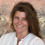 Jennifer Kircher Herman's hair cascades down her back. She smiles at the camera in a white linen shirt, in front of a blurry abstract tan background. 