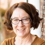 Author photo: Jody Hobbs Hesler has plastic frame rounded glasses, ear length fluffy brown hair and a yellow ochre top. She smiles in the sunlight.