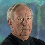 Artist photo: Brian McPartlon looks at the camera from in front of a blue, abstract background. He wears a black shirt, and his gray/white hair reflects the light.