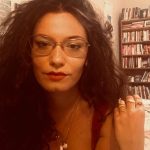 Rachel Salvini sits in front of a bookshelf, in glasses and a tank top. Her hand rests on her shoulder and she gazes into the camera, her dark hair taking up most of the frame.