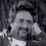 Author photo of Virgil Suárez. In black and white, he sits with a smile on his face, ringed by a goatee. Hand on his cheek, and wire frame glasses before warm eyes. His hair is fluffy and he wears an unbuttoned shirt over a white tee shirt. Blurry gray-black background.