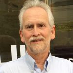 Author photo of George Franklin, white man with white/gray hair in a pale blue shirt, he looks at the camera in front of a dark wood wall.