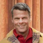 Michael Anderson smiles in a red shirt under a brown coat. His short blond hair is combed smooth and he stands before an orange curtain.