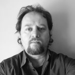 Edward Lee, photographed in silhouette in a dark room with wood and glass doors. Behind him, through the doorway is a brighter room with light wood and white paint.