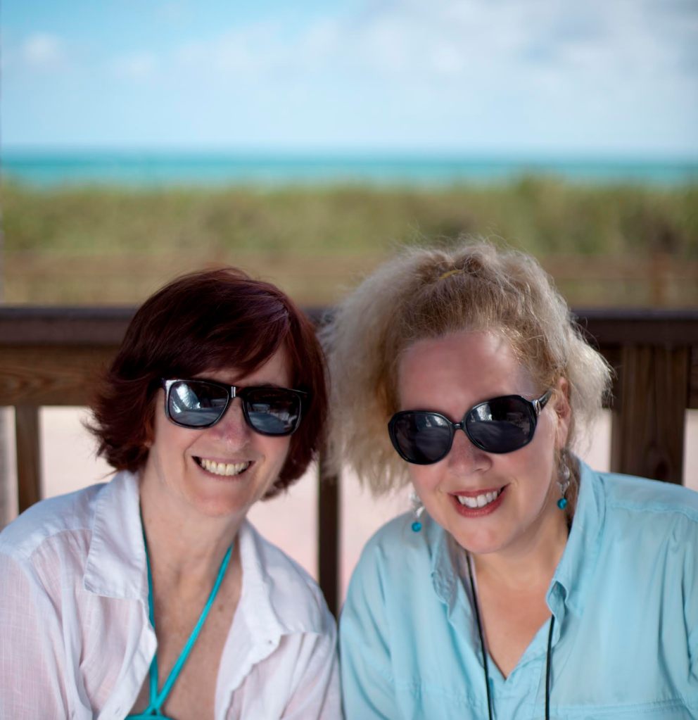 Maureen Seaton and Denise Duhamel smile at the camera, shoulders touching. Both wear sunglasses. They're in front of a wooden railing, then grass, then the beach. Denise wears light blue and Maureen white, with a light blue lanyard. They look happy together.