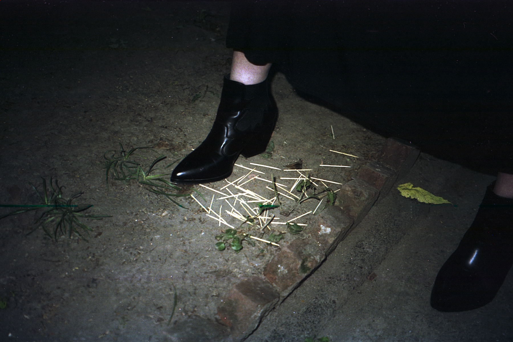 Shiny black boot stepping on scattered toothpicks and weeds on the dirt ground. A line of bricks between the two feet. Lit from above by flashlight. 